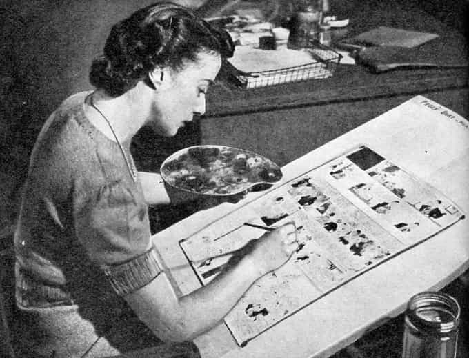 Image shows a female colorist in the 1940s wearing a sweater and necklace, holding a palette of watercolors in her left hand, and painting a full-page cartoon on a large drawing board with her right (black and white).